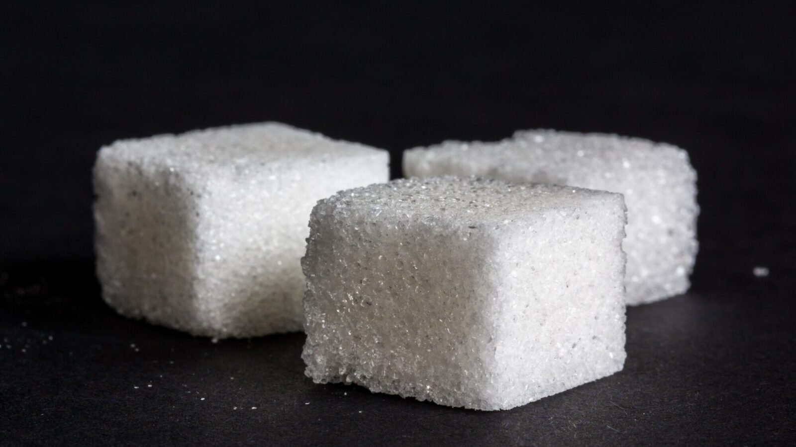 Drei weiße Zuckerwürfel liegen nebeneinander vor dunklem Hintergrund in Nahaufnahme